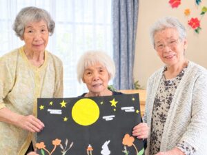メイン（男性が貼り付け中）：黒画用紙にお月見の飾りを貼り付けるご利用者様 お月見の創作活動 大田区上池台のデイサービス ファミリーケア心の家 上池台
