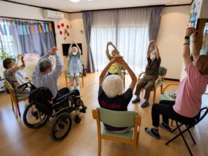 椅子に座って両手を上げ棒体操に取り組むご利用者様 大田区上池台のデイサービス ファミリーケア心の家 上池台